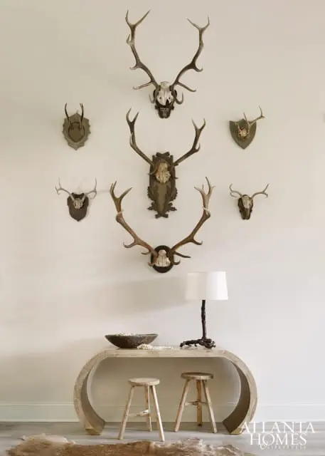 Wall decorated with mounted deer antlers and skulls above a light wood console table with two stools, a bowl, and a lamp.