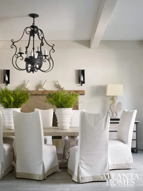 Dining room with a rustic wooden table and slipcovered chairs, featuring a black wrought iron chandelier, a stone fireplace with decorative birds, and potted ferns.