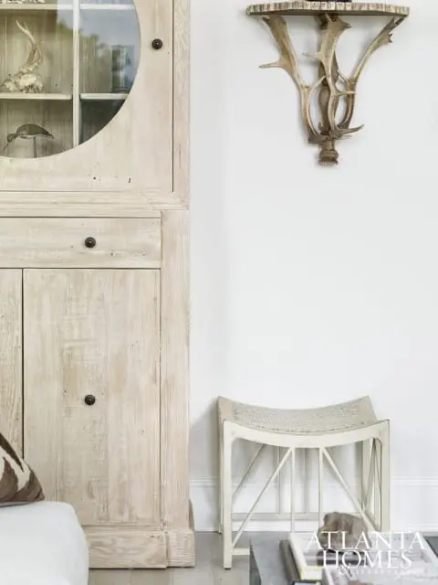 Light wood hutch with glass door displaying taxidermy and decorative objects, next to a white bench with woven seat and antler wall sconce.