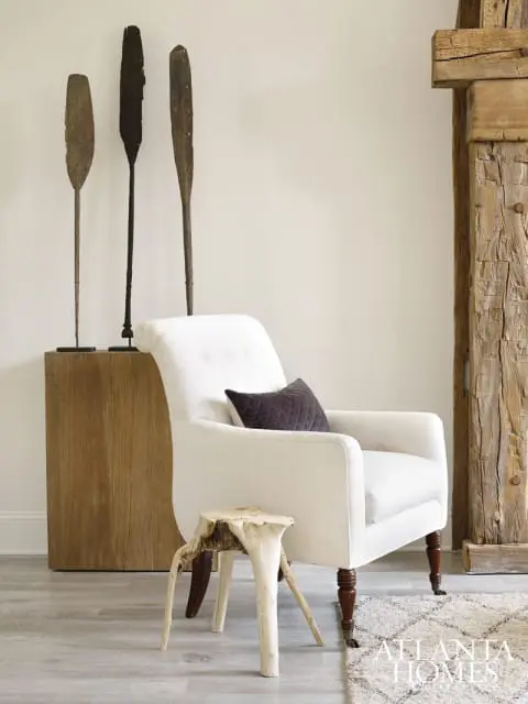 Cream upholstered armchair with a dark quilted pillow, next to a rustic wooden side table and a display of three wooden oars on a wooden pedestal.