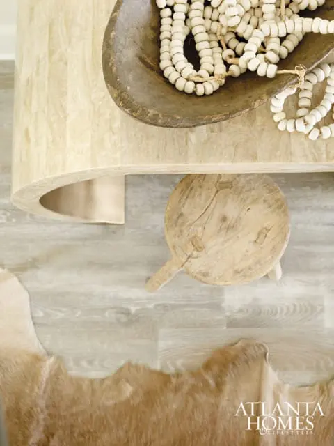 Light wood console table with curved base, vintage wooden bowl with white beads, small wooden stool, and tan hide rug on wood-look floor.