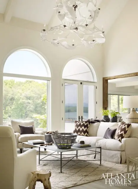 Living room with vaulted ceiling, large arched windows overlooking trees and a lake, a white linen sofa with patterned pillows, and a metal coffee table with a distressed metal bowl.