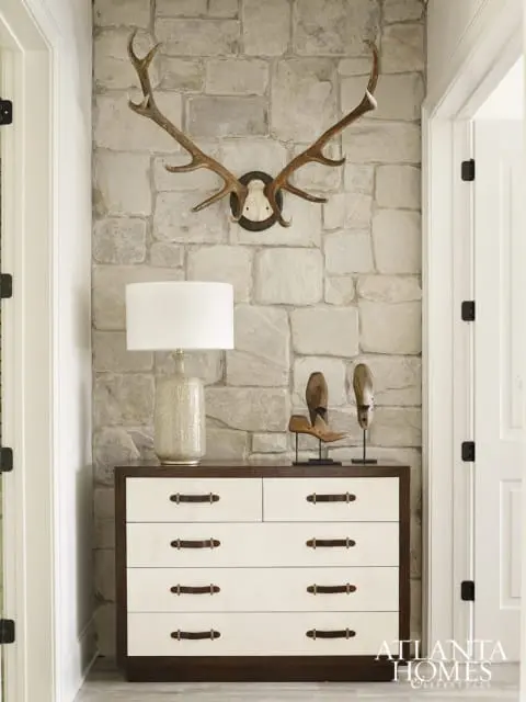 Hallway with stone accent wall featuring mounted deer antlers, a textured ceramic lamp, and a white dresser with dark wood trim and leather drawer pulls.