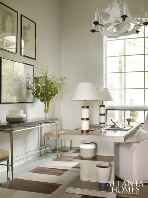 Neutral-toned office with a large window, antler chandelier, framed botanical prints, and a desk with two lamps.