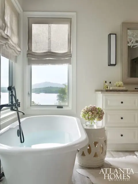 White clawfoot bathtub with black faucet, linen roman shades, and a white ceramic garden stool with hydrangeas. A white dresser with black knobs and a decorative mirror are visible.