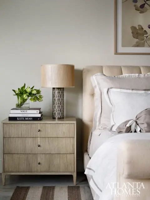 Bedroom with a light beige tufted headboard, stacked books and a patterned lamp on a ribbed wood nightstand, and a striped rug.