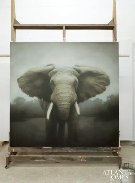 Painting of an elephant with large ears and tusks, set against a misty background of trees. The painting is on an easel in a studio.