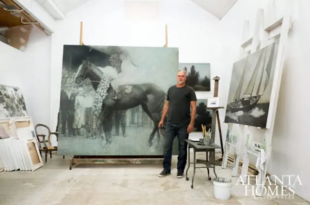 Man standing in an art studio next to a large painting of a horse and a painting of a sailboat.