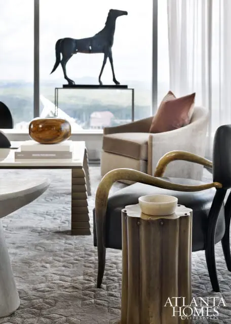 Living room with a dark metal horse sculpture on a pedestal, a wooden bowl on a stack of books, and upholstered chairs with brass accents.