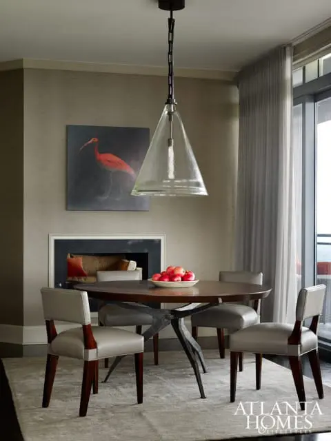 Dining room with round wood table, four grey upholstered chairs, fireplace with artwork of a red bird, and a glass pendant light.