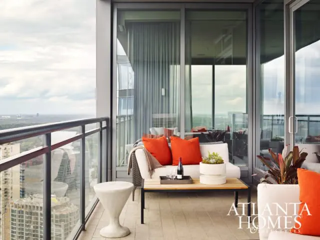Modern balcony with white sofa, orange accent pillows, wooden coffee table, and concrete stool overlooking a city skyline.