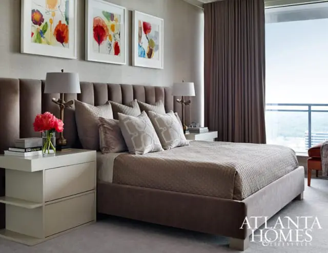 Bedroom with a plush brown headboard, neutral bedding, and three framed floral art pieces above the bed. A modern white nightstand holds a lamp and flowers.
