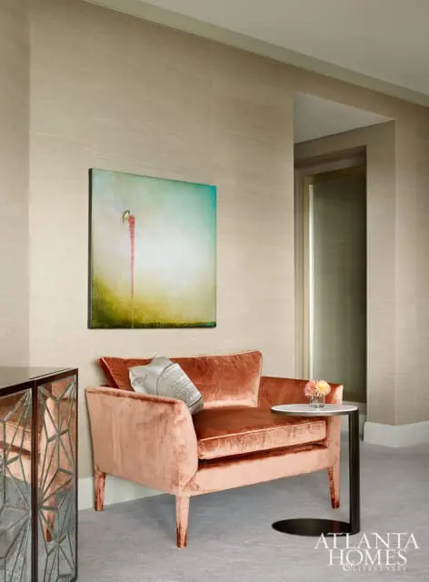 Peach velvet armchair with a silver patterned pillow, next to a small round side table with a vase of flowers. A textured grasscloth wallpaper covers the walls.