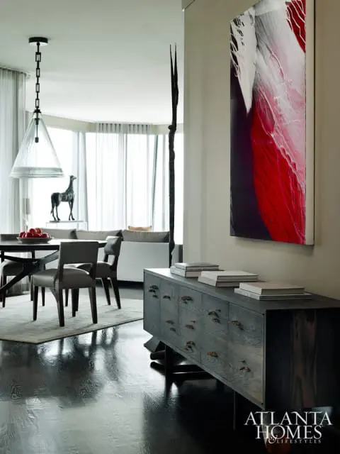 Dining area with dark wood table and chairs, a gray rug, and a dark wood credenza topped with books. A large abstract painting with red and white accents hangs on the wall.