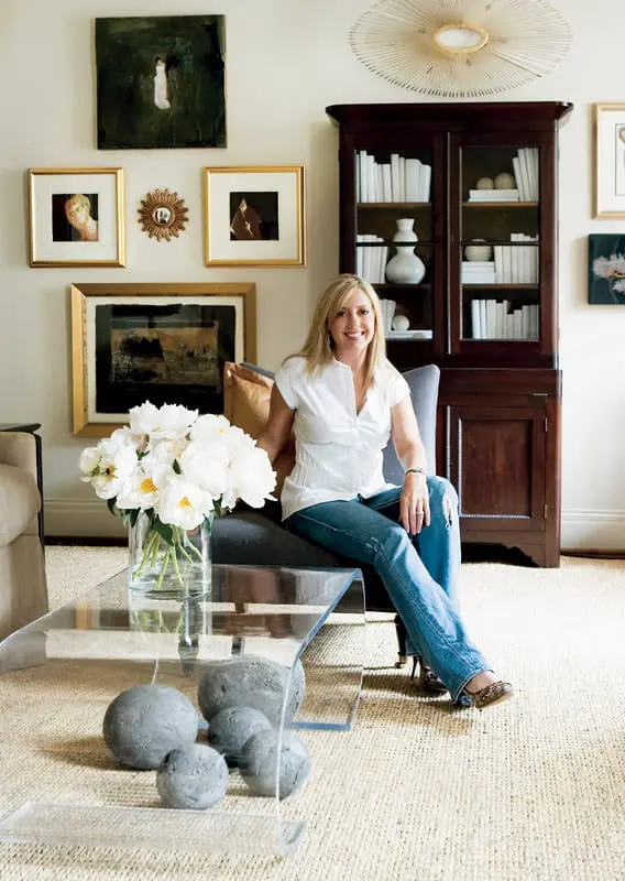 Woman in white blouse and jeans sitting in a charcoal armchair in a living room with a dark wood cabinet filled with books and a clear acrylic coffee table with white peonies and gray spheres.