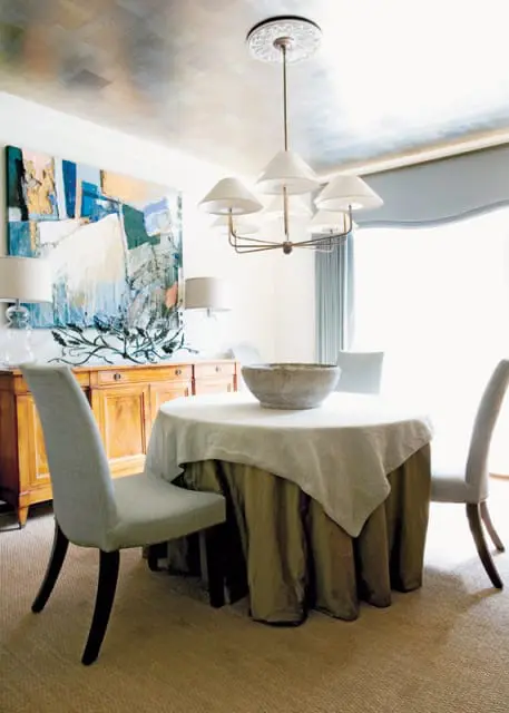 Dining room with a round table covered in a light linen tablecloth, a large stone bowl, and upholstered chairs. A wooden buffet with artwork hangs on the wall, and the ceiling features a metallic finish.
