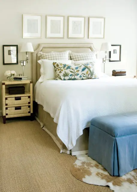 Bedroom with a tufted headboard, white bedding, and a patterned lumbar pillow. A woven nightstand and a blue upholstered bench are visible.