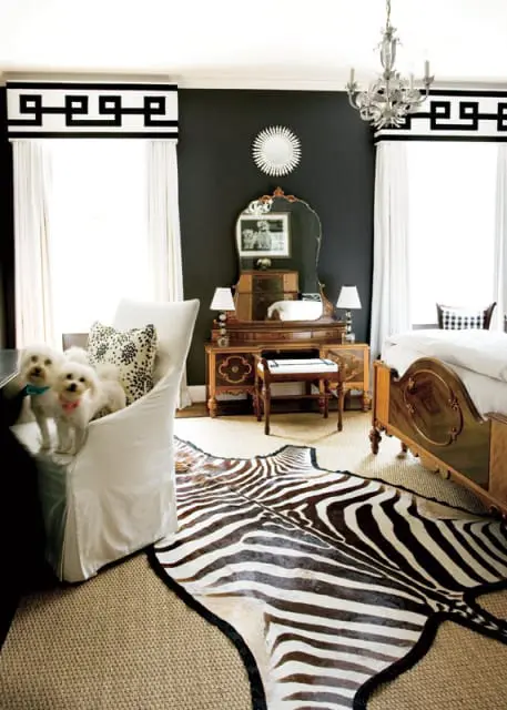 Dark gray bedroom with white curtains, Greek key valances, a vintage vanity with a mirror, a zebra print rug, and two white dogs on a slipcovered chair.