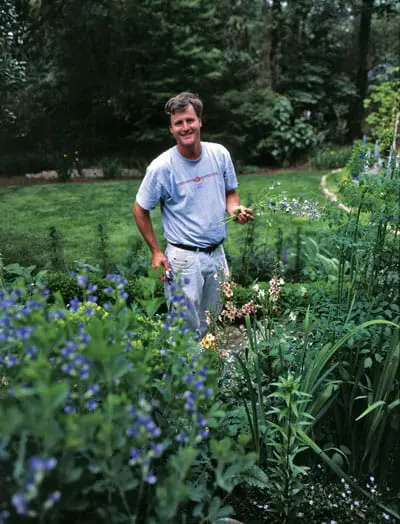 Man in a t-shirt and jeans standing in a lush garden, holding a small bunch of flowers and pruning shears.