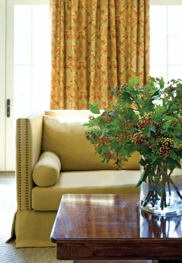 Yellow sofa with nailhead trim and bolster pillow, in front of a patterned curtain. A wooden table holds a vase of greenery with red and yellow berries.