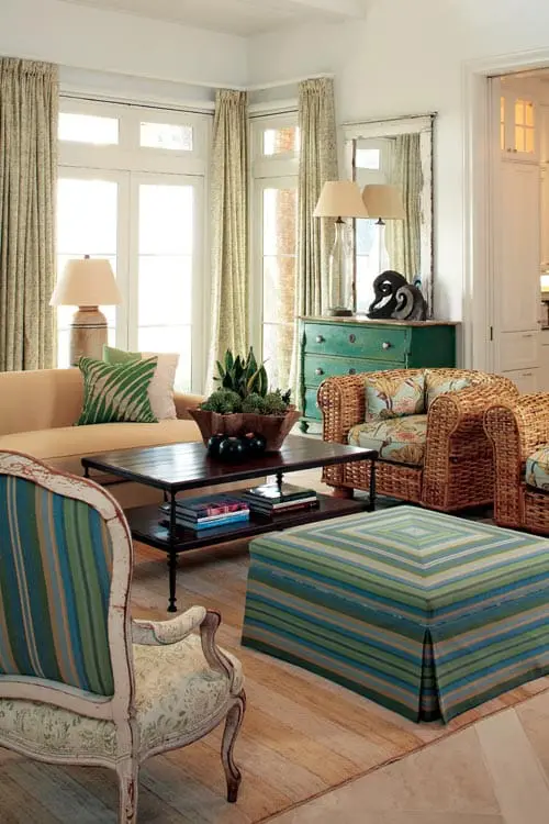 Living room with a light yellow sofa, wicker armchairs, a dark wood coffee table, and a striped ottoman. Green dresser with mirror in background.