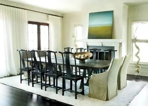 Formal dining room with a long dark wood table and twelve dark wood chairs, a large white rug, and a fireplace with abstract art above it.