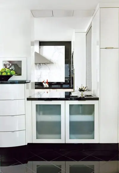 Modern white kitchen with black granite countertops, stainless steel range hood, and frosted glass cabinet doors. A bowl of green apples sits on a curved cabinet to the left.