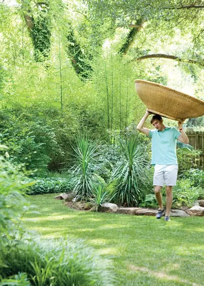 Man carrying a large woven basket on his head walks across a lush green lawn surrounded by bamboo, yucca plants, and trees.