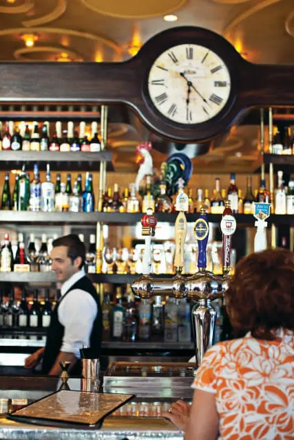 Bar interior with a bartender in a black vest and white shirt, shelves stocked with bottles, beer taps, and a large clock above the bar.