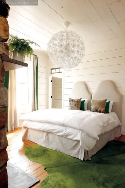 Bedroom with white shiplap walls, a large white dandelion-shaped chandelier, a stone fireplace, and a bed with linen headboards and white bedding. A bright green rug covers part of the wood floor.