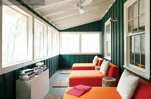 Sunroom with dark green paneled walls, white ceiling, and floor-to-ceiling windows. Orange chaise lounges with white pillows line the room, with small wood stump side tables.