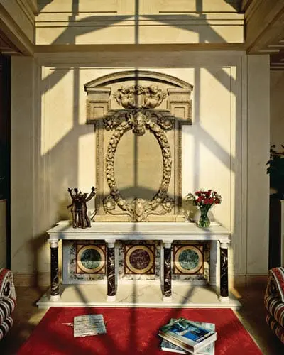 Interior hallway with a marble console table featuring inlaid circular medallions, topped with a sculpture and vase of flowers, beneath an ornate carved wall niche.