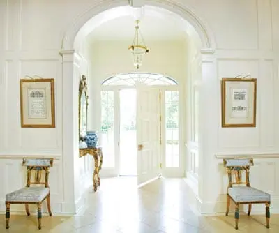 White entryway with paneled walls, arched doorway, double doors, antique chairs, and a console table with a mirror and vase.