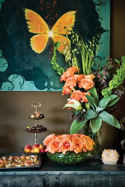 A table setting with appetizers, a tiered fruit stand, and floral arrangements, beneath a painting of an orange butterfly on a dark teal background.