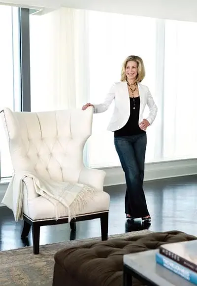 Woman in white blazer and jeans standing next to a tufted white armchair in a well-lit room.