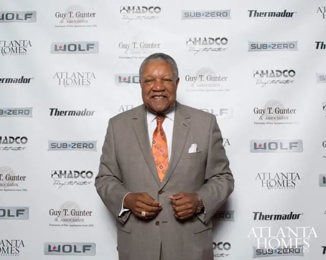 Man in a tan suit and orange patterned tie smiles in front of a backdrop with repeating logos for Atlanta Homes & Lifestyles, Wolf, Sub-Zero, Thermador, AHADCO, and Guy T. Gunter & Associates.