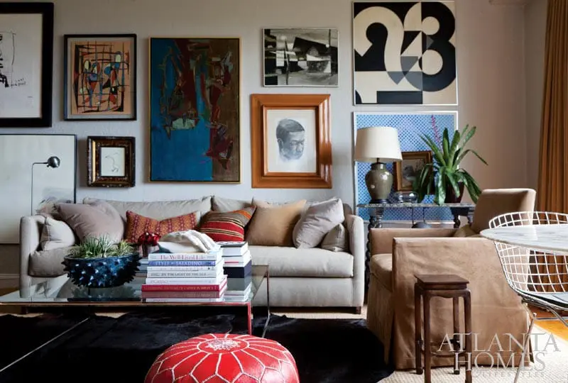 Living room with a neutral sofa, eclectic artwork, glass coffee table with books, and a red pouf.