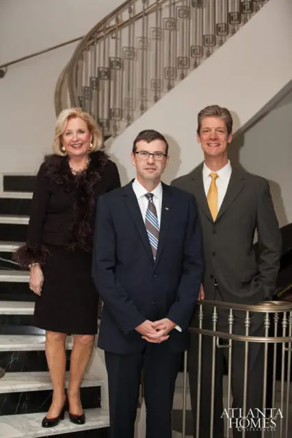 A woman in a dark brown dress with fur trim stands on a marble staircase next to two men in suits. The man in the center wears a navy suit and a striped tie. The man on the right wears a charcoal suit and a yellow tie.