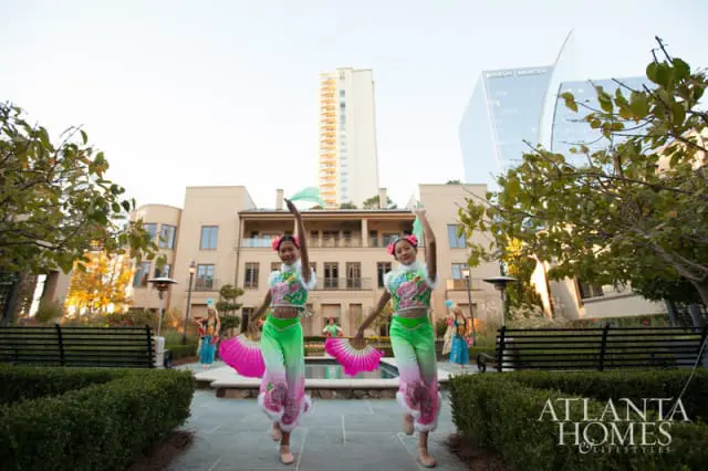 Two young women in traditional Chinese dance costumes with pink and green fans perform in a courtyard with a building and skyscrapers in the background.