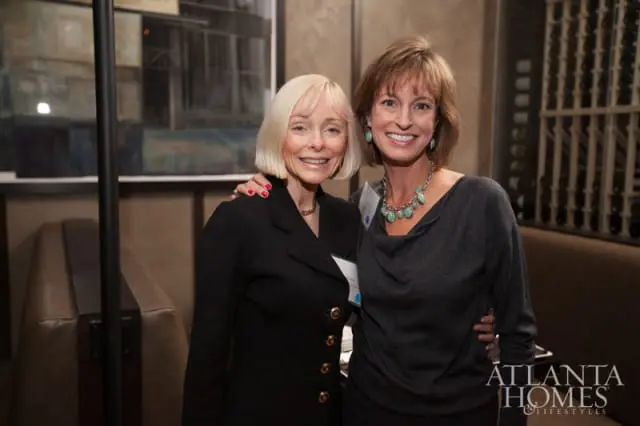 Two women smiling at a broker event, one with blonde bob haircut wearing a black blazer, the other with brown hair wearing a gray cowl-neck top.