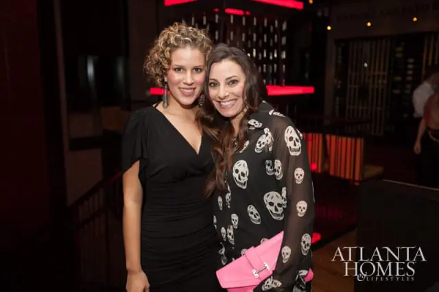 Two women smiling at the camera at an event. The woman on the left wears a black dress and the woman on the right wears a black shirt with a skull print and holds a pink clutch.