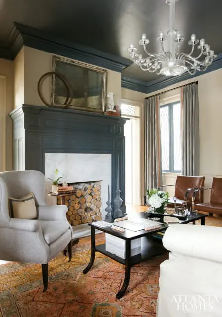 Gray armchair in living room with dark blue fireplace, marble surround, stacked logs, and ornate chandelier on dark ceiling.