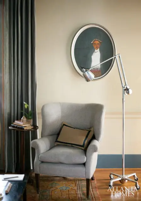 Gray upholstered wingback chair with a tan pillow, next to a small side table with books and a plant. A silver floor lamp stands nearby, and a framed portrait hangs on the wall.