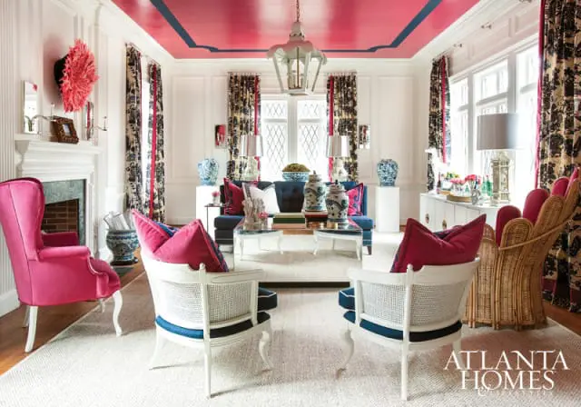 Living room with a bright pink ceiling with navy blue trim, white paneled walls, patterned curtains, and a mix of furniture including a navy sofa, white cane chairs with navy cushions, and a pink velvet armchair.