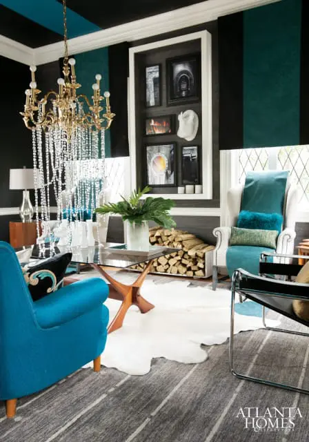Living room with teal and black striped wallpaper, a gold crystal chandelier, a glass coffee table, a white cowhide rug, and a teal armchair.