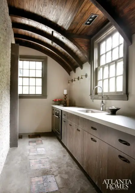 Kitchen with vaulted wood ceiling, concrete floor with wood inlay path, light stone wall, and dark wood cabinetry with white countertop.