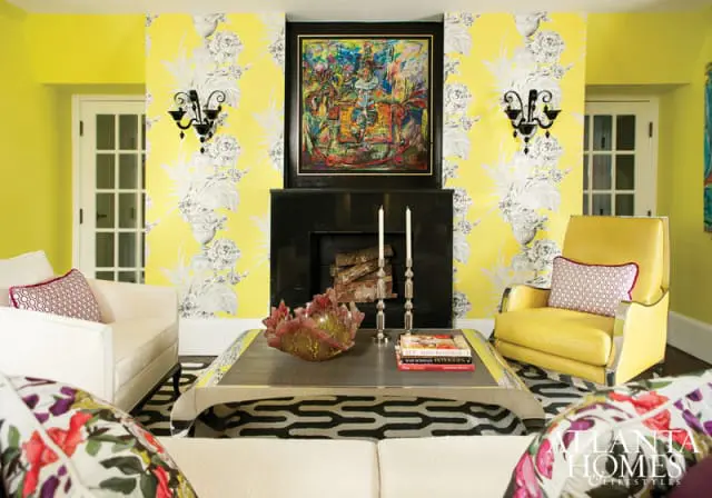 Living room with bright yellow walls and floral wallpaper, featuring a black fireplace, a colorful abstract painting, white sofas, a yellow armchair, and a patterned rug.