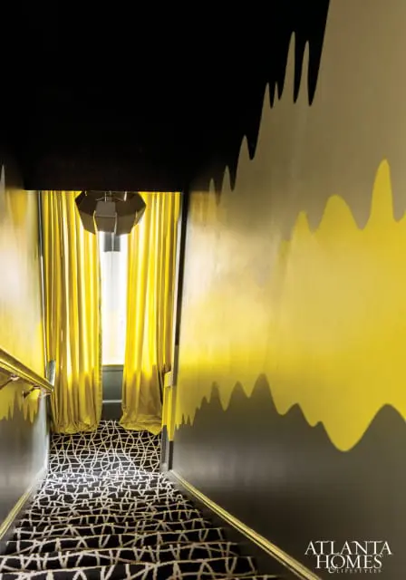Dark gray hallway with a black ceiling and a yellow and gray abstract wave mural. A black and white geometric patterned carpet covers the stairs, with bright yellow curtains framing a window at the top of the stairs.