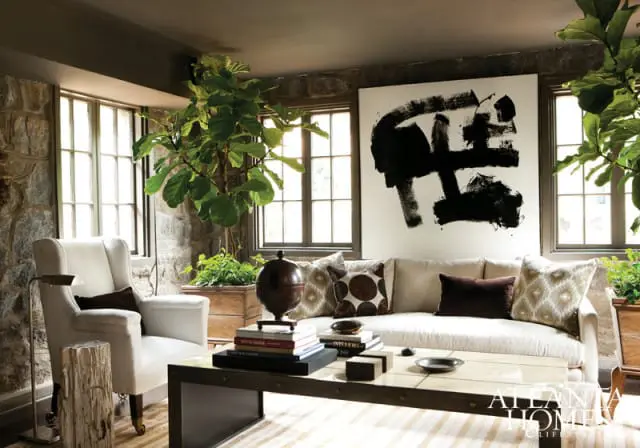 Living room with stone walls, a white wingback chair, a neutral sofa with patterned pillows, a large abstract painting, and a fiddle-leaf fig tree.