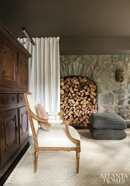 Living room with a vintage wooden cabinet, ornate gold-trimmed chair with a pink pillow, stacked grey poufs, and a stone fireplace filled with firewood.